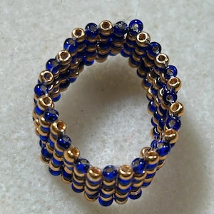 K&ouml;nnte beinhalten: Handgefertigter Ring aus goldenen und tiefblauen Perlen. Der Ring besteht aus mehreren Reihen kleiner Perlen, wodurch ein strukturiertes Aussehen entsteht. Ein Schmuckst&uuml;ck, das zu jedem Outfit passt.