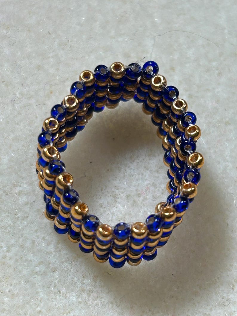 K&ouml;nnte beinhalten: Handgefertigter Ring aus goldenen und tiefblauen Perlen. Der Ring besteht aus mehreren Reihen kleiner Perlen, wodurch ein strukturiertes Aussehen entsteht. Ein Schmuckst&uuml;ck, das zu jedem Outfit passt.