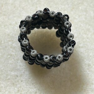 K&ouml;nnte beinhalten: Handgefertigter Perlenring mit abwechselnden Reihen aus schwarzen und grauen Perlen. Der Ring hat ein mehrschichtiges Design, das eine strukturierte Oberfl&auml;che zeigt. Die Perlen sind klein und rund, was ein einzigartiges Accessoire ergibt.