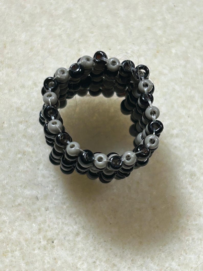 K&ouml;nnte beinhalten: Handgefertigter Perlenring mit abwechselnden Reihen aus schwarzen und grauen Perlen. Der Ring hat ein mehrschichtiges Design, das eine strukturierte Oberfl&auml;che zeigt. Die Perlen sind klein und rund, was ein einzigartiges Accessoire ergibt.