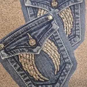 May include: Close-up of two denim jean pockets. The pockets are a medium blue wash with white stitching. Each pocket has a decorative wing design made of small, sparkling rhinestones. Silver-colored buttons and rivets add detail.