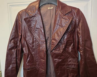 May include: A burgundy leather jacket with a notched collar and two front pockets hangs on a hanger. The jacket has a classic, tailored design with a button closure and a satin lining. The leather appears worn, giving it a vintage look.