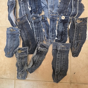May include: A collection of denim jean waistbands and flys, cut from various pairs of jeans. The pieces are in shades of blue, with visible buttons, zippers, and stitching details. The arrangement suggests a craft or upcycling project.
