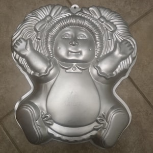 May include: A silver-colored metal cake pan shaped like a doll with yarn-style hair in pigtails, a round face with dimples, raised arms, and legs tucked inward. The mold features detailed impressions for the facial features, clothing, and hair bows.