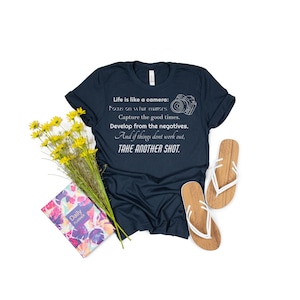 May include: A navy blue t-shirt with a white graphic of a camera and the text "Life is like a camera: Focus on what matters. Capture the good times. Develop from the negatives. And if things don't work out, TAKE ANOTHER SHOT."