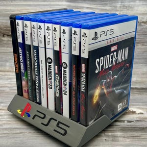 Può includere: Un supporto grigio per giochi PS5 con una collezione di custodie di giochi blu e nere. Il supporto presenta il logo PS5 e contiene giochi come Spider-Man: Miles Morales e Far Cry 6. I giochi sono esposti verticalmente.