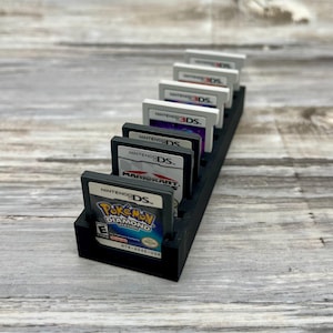 Puede incluir: Un soporte negro y rectangular para cartuchos de juegos, lleno de cartuchos de juegos de Nintendo DS y 3DS. Los cartuchos muestran títulos de juegos y el logotipo de Nintendo. El soporte es de material oscuro.