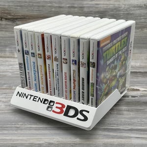 May include: A white plastic display stand featuring the Nintendo 3DS logo in black and red text, holding a row of ten Nintendo 3DS video game cases. The titles include Teenage Mutant Ninja Turtles: Danger of the Ooze, Mario Kart 7, and various Pokémon games.