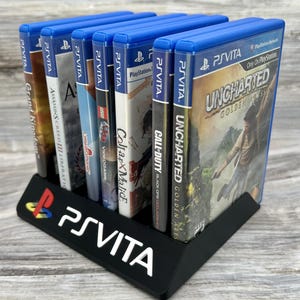 以下が含まれることがあります： 白いPS Vitaロゴが入った黒いプラスチック製のディスプレイスタンド。PlayStation Vita用ゲームソフトのケースが7つ収納されています。確認できるゲームタイトルには、『アンチャーテッド 地図なき冒険の始まり』、『コール オブ デューティ ブラックオプス デクラシファイド』、『グランド・キングダム』などがあります。