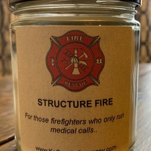 May include: A glass jar candle with a brown label featuring a red firefighter's cross with the words "FIRE RESCUE" and the text "STRUCTURE FIRE For those firefighters who only run medical calls... www.KcCandleCompany.com"