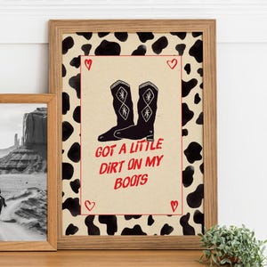 Puede incluir: Impresión enmarcada con botas de vaquero negras y el texto "Got a little dirt on my boots" en rojo. El fondo presenta un diseño de vaca. El marco de madera añade un toque rústico.