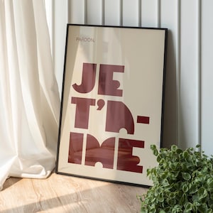 May include: A framed art print with the words "JE T'IME" in bold, burgundy letters on a cream background. The print is in a black frame and leans against a white wall. A small green plant is in the lower right corner.
