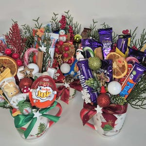 Puede incluir: Dos tazas navideñas llenas de golosinas festivas. Cada taza está repleta de caramelos, chocolates, adornos y vegetación. Las tazas están decoradas con motivos navideños y atadas con lazos, creando un regalo alegre.