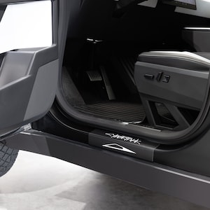 May include: A black car interior with a gray door open, revealing a black floor mat with the word "Shark" printed on it. The car has a black leather seat and a silver metal pedal.