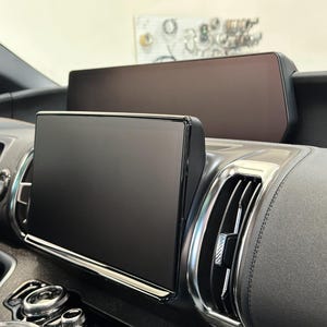 May include: A car interior with two black screens, one above the other. The top screen is larger than the bottom screen. The screens are mounted on a black dashboard. The dashboard is made of a textured material. The screens are off and the reflection of the surrounding area is visible on the screens.