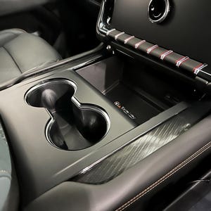 May include: Close-up of a car's interior showing a black leather center console with two cup holders, a storage compartment, and a carbon fiber trim. The console has a red and white stripe design.