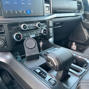 May include: A black phone mount attached to the center console of a car. The mount has a circular magnetic surface. The car has a black leather interior and a gear shifter.