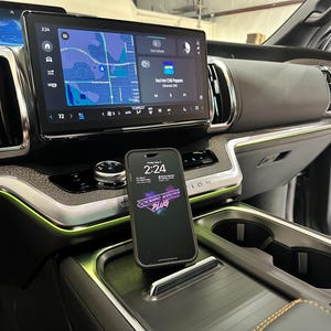 May include: Interior view of a vehicle's dashboard, featuring a large touchscreen displaying a map and music information. A smartphone with the time 2:24 and a graphic is in the foreground. The dashboard has a modern design with ambient lighting.
