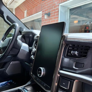 May include: A black car interior with a large touchscreen display, a silver knob, and a 12V and AC 120V outlet. The screen is displaying a dark background.