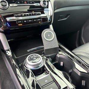 May include: A black car interior with a wireless charging pad in the center console. The charging pad has a lightning bolt icon on it. The console also has a gear shifter and two cup holders.