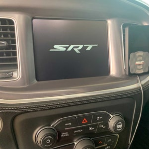 May include: A close-up of a car's dashboard with a large screen displaying the word "SRT". The screen is surrounded by buttons and controls, including a volume knob, drive mode selector, launch control button, and climate control buttons.
