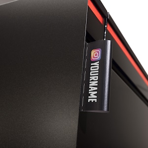 May include: Black computer case with a red stripe and a black tag hanging from the top. The tag has a colorful Instagram logo and the text "YOURNAME" printed on it.
