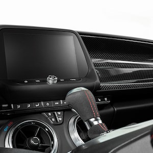 以下が含まれることがあります： 黒いダッシュボードと大きなタッチスクリーンディスプレイを備えた車のインテリア。ダッシュボードはカーボンファイバートリムと、赤いステッチが施された黒い革製のシフトレバーを備えています。