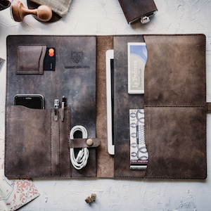 May include: A brown leather portfolio with multiple pockets and compartments. The portfolio is open and has a tablet, phone, pens, and a charging cable inside. The portfolio has the text "WAXED BROWN" embossed on the front.