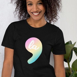 Pode incluir: T-shirt preto com um gráfico de vírgula colorido. A vírgula é preenchida com cores pastel e as letras "la" estão escritas dentro. A modelo está usando jeans azuis.