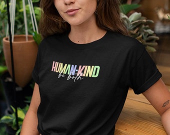 Puede incluir: Una mujer lleva una camiseta negra con el texto "HUMAN-KIND be both" en colores arcoíris. Está sentada en una superficie de madera con plantas en el fondo.