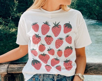 Puede incluir: Camiseta blanca con un patrón repetido de fresas rojas con tallos verdes. La camiseta se combina con pantalones cortos de mezclilla. La imagen se toma al aire libre, con un río y árboles al fondo.