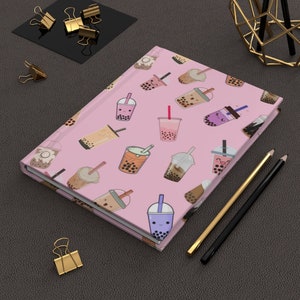 May include: A pink notebook with a pattern of colorful bubble tea cups with smiling faces. The notebook is closed and lying on a gray surface with two pencils and a gold paper clip.