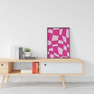 Op de afbeelding: Een ingelijst afdruk van een abstract ontwerp in roze en wit met zwarte lijnen. De afdruk hangt aan een witte muur boven een houten plank met boeken en een plant in een pot.
