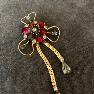May include: A vintage brooch featuring a floral design with red and clear gemstones. The brooch has a gold-tone metal frame and chain accents, with teardrop-shaped clear stones dangling from the bottom.