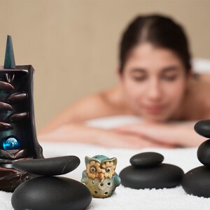 May include: A ceramic incense burner with a blue and brown design, a small ceramic owl figurine, and a stack of black stones on a white surface.
