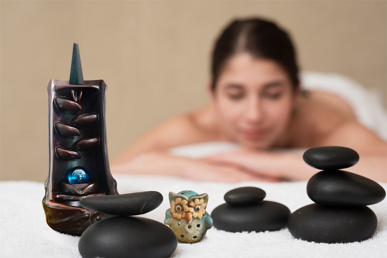 May include: A ceramic incense burner with a blue and brown design, a small ceramic owl figurine, and a stack of black stones on a white surface.