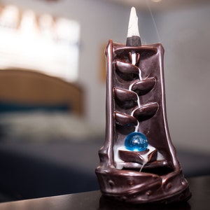 May include: A brown ceramic incense burner with a blue glass ball in the center. Smoke flows from the top of the burner, creating a waterfall effect.
