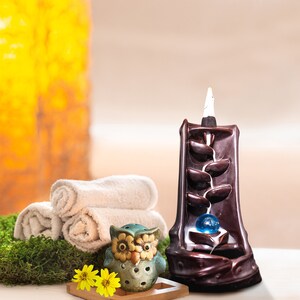 May include: A brown ceramic incense burner shaped like a waterfall with a blue glass ball in the center. A small owl incense burner is in the foreground. The incense burner is on a white surface with a green moss background.