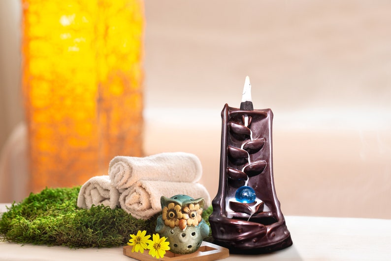 May include: A brown ceramic incense burner shaped like a waterfall with a blue glass ball in the center. A small owl incense burner is in the foreground. The incense burner is on a white surface with a green moss background.