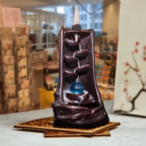 May include: A brown ceramic incense burner shaped like a waterfall with a blue glass ball in the center. The incense burner is on a wooden base.