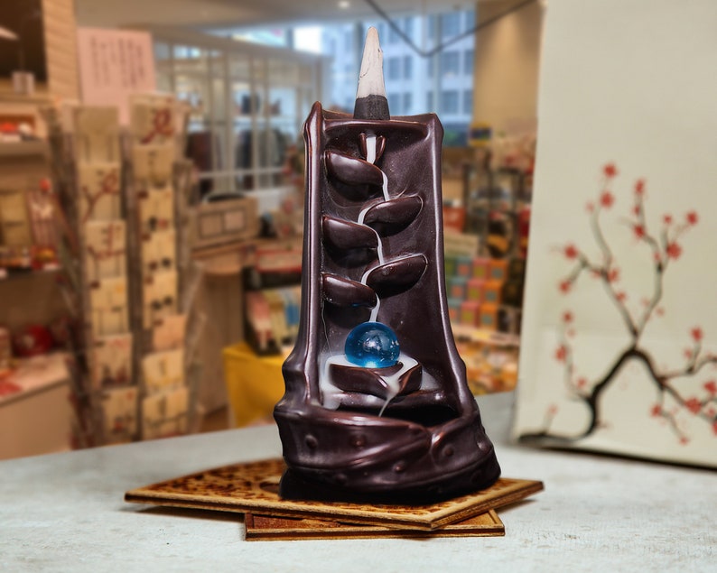 May include: A brown ceramic incense burner shaped like a waterfall with a blue glass ball in the center. The incense burner is on a wooden base.