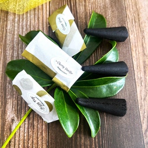 May include: Three black incense cones on a green leaf with three small packages of incense. The packages are labeled "Ocean Mist", "Cherry Vanilla", and "Dolce Melya".
