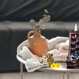 May include: A brown ceramic incense burner with a cascading design and a blue glass ball in the center. The burner is on a white table with a knitted throw, a small ceramic owl figurine, and yellow flowers.