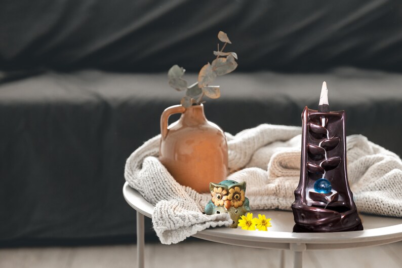 May include: A brown ceramic incense burner with a cascading design and a blue glass ball in the center. The burner is on a white table with a knitted throw, a small ceramic owl figurine, and yellow flowers.