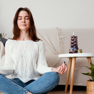 May include: A woman sits cross-legged on the floor with her eyes closed, meditating. She is wearing a white sweater and blue jeans. A small table with a brown incense burner and a green plant in a wicker basket is in the background.
