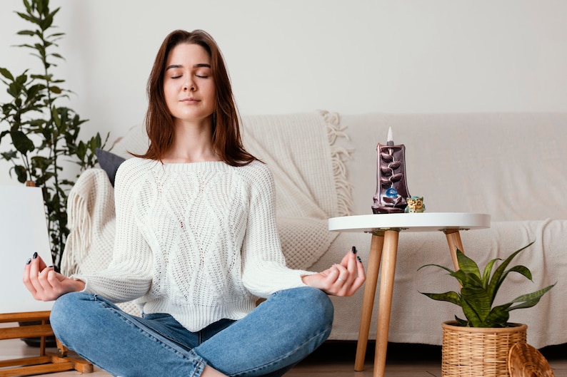 May include: A woman sits cross-legged on the floor with her eyes closed, meditating. She is wearing a white sweater and blue jeans. A small table with a brown incense burner and a green plant in a wicker basket is in the background.