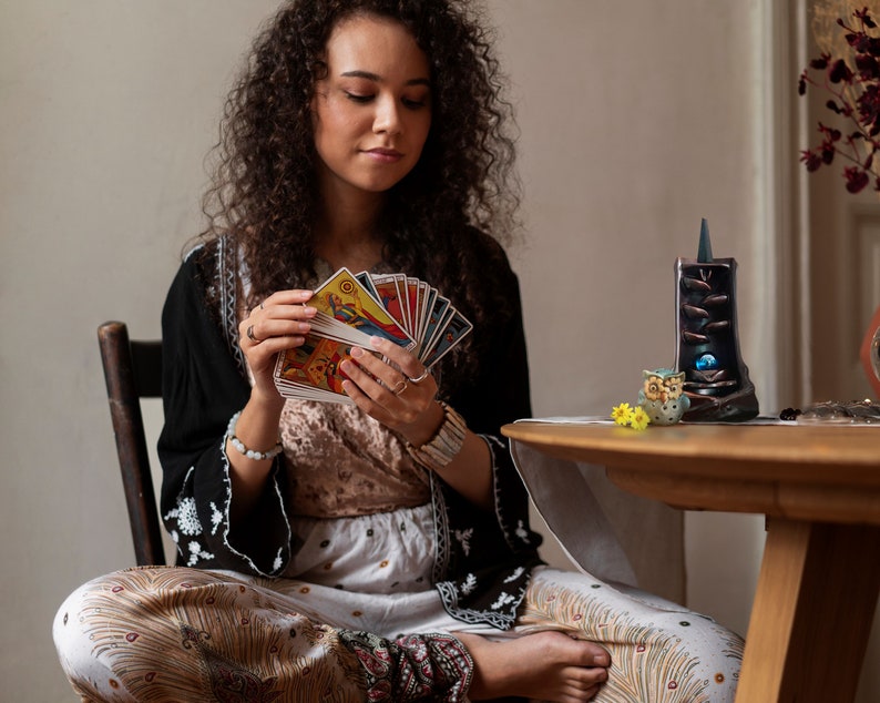 May include: A woman sits in a chair, holding a spread of tarot cards in her hands. She is wearing a long-sleeved black shirt with white embroidery and a white and brown patterned skirt. A small ceramic owl figurine and a black incense burner are on a wooden table in front of her.
