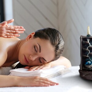 May include: A woman receiving a massage on a white table. A black ceramic incense burner with a blue glass orb and a white incense cone is on the table next to her.