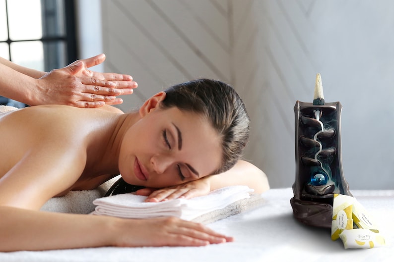 May include: A woman receiving a massage on a white table. A black ceramic incense burner with a blue glass orb and a white incense cone is on the table next to her.