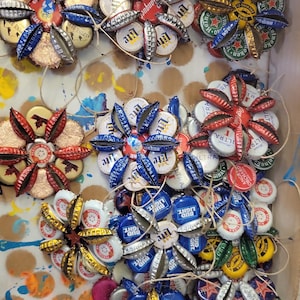 Peut inclure: Ornements floraux faits à la main à partir de capsules de bouteilles de bière. Chaque fleur présente une palette de couleurs unique, avec des capsules bleues, rouges, dorées et argentées disposées en cercle. Les ornements sont reliés par de la ficelle.